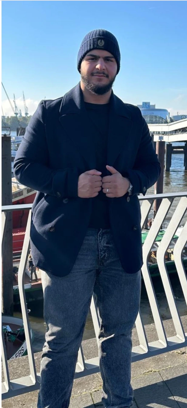Deniz steht mit Mütze und dunklem Mantel am Hafen.